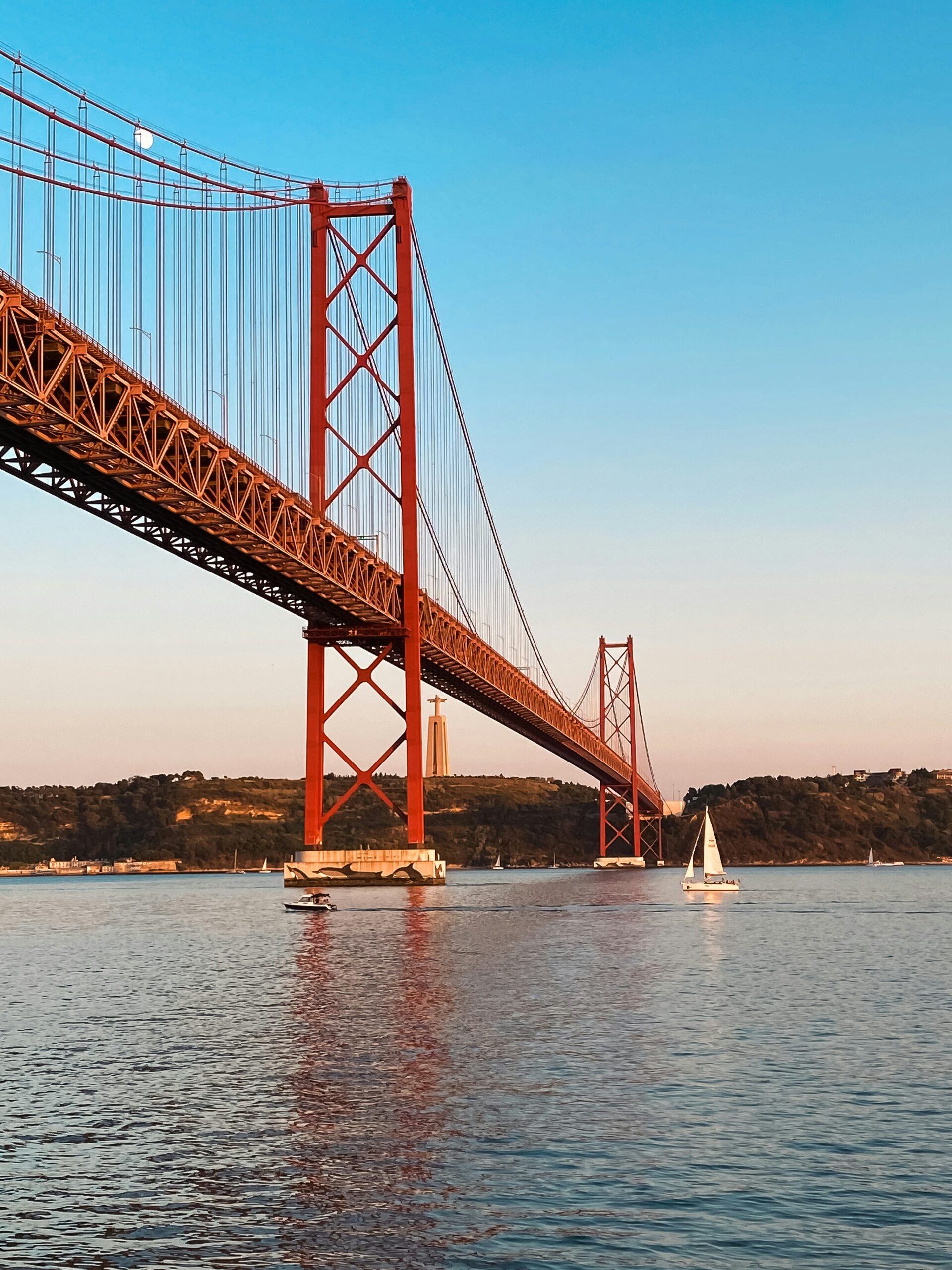 Lisbon's 25 de Abril Bridge, beneath a beautiful sunset, sailing boats grace the waters below.