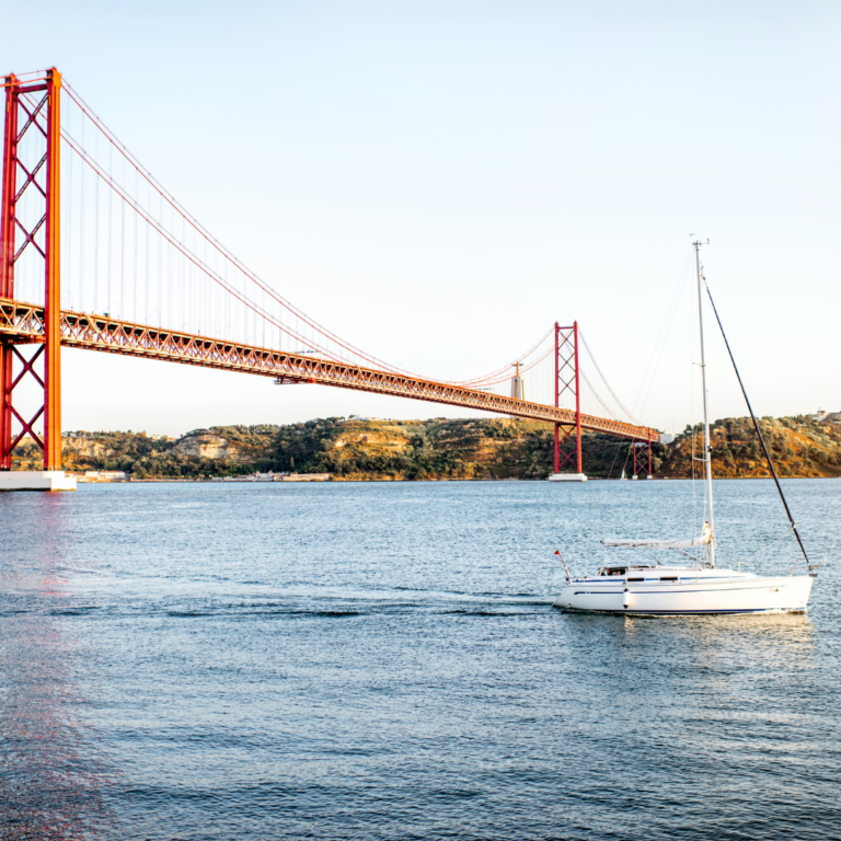 A beautiful white sailing boat gliding beneath Lisbon's iconic red bridge.
