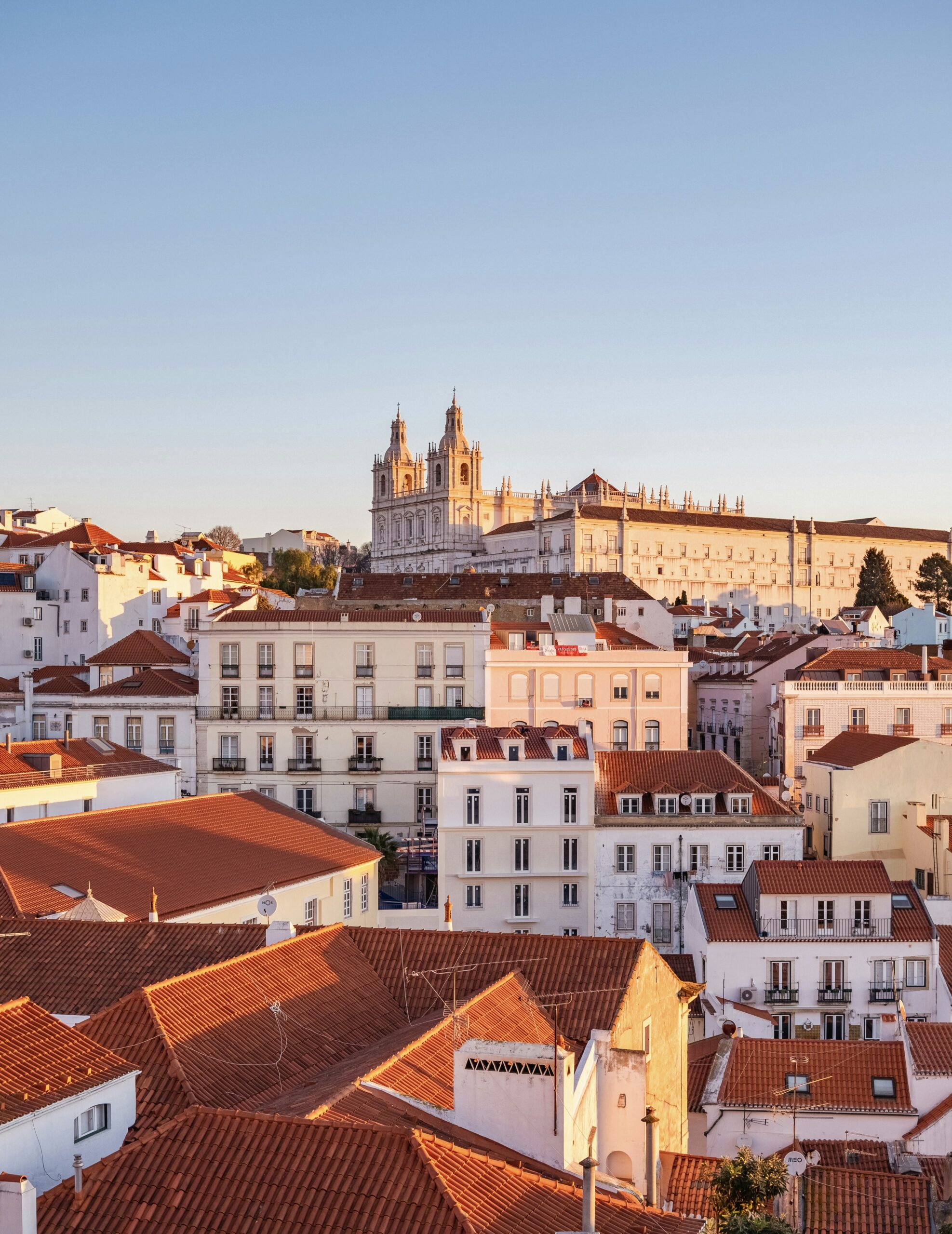 Lisbon aerial view: Red rooftops, Jerónimos Monastery backdrop, bathed in summer evening glow.